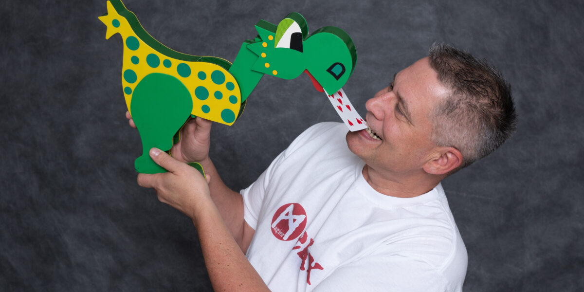 Ein Mann in einem weißen T-Shirt hält eine grün-gelb gepunktete Dinosaurierpuppe, die in der Nähe seines Mundes auf einer Spielkarte herumzukauen scheint.