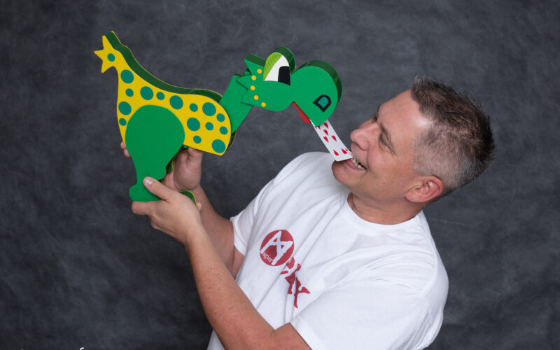 Ein Mann in einem weißen T-Shirt hält eine grün-gelb gepunktete Dinosaurierpuppe, die in der Nähe seines Mundes auf einer Spielkarte herumzukauen scheint.