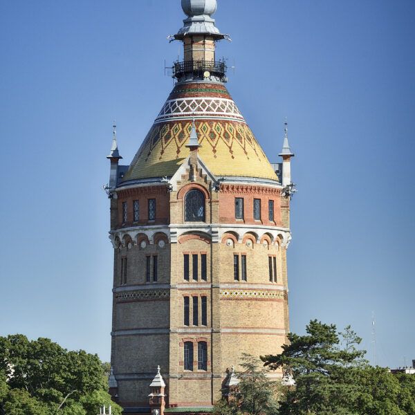 Ein hoher Ziegelstein-Wasserturm mit einem gelben Ziegeldach und kunstvollen Details, umgeben von grünen Bäumen unter einem klaren blauen Himmel.