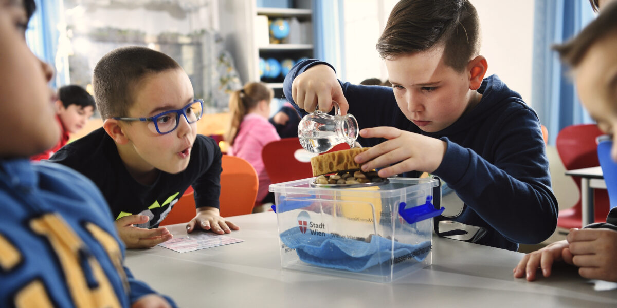 Zwei Jungen führen an einem Tisch im Klassenzimmer ein wissenschaftliches Experiment mit Wasser und Schwämmen durch, während andere Kinder zusehen.