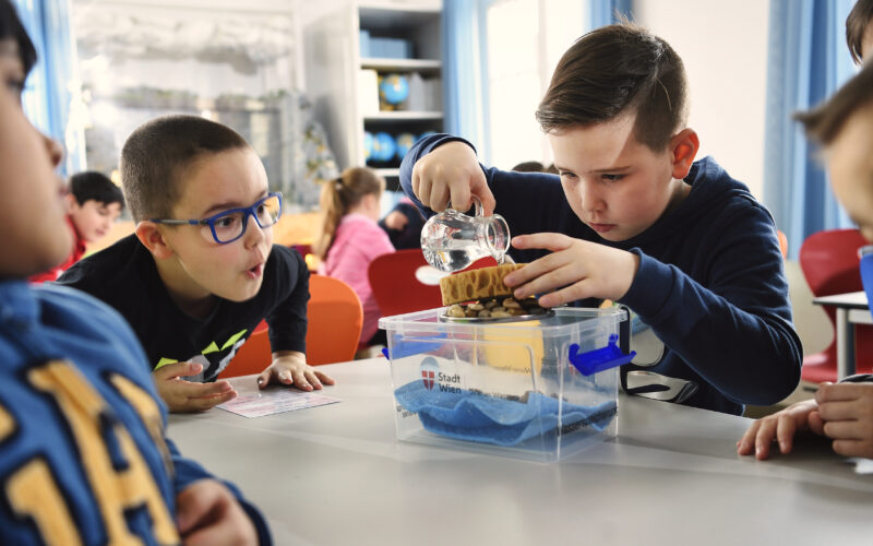 Zwei Jungen führen an einem Tisch im Klassenzimmer ein wissenschaftliches Experiment mit Wasser und Schwämmen durch, während andere Kinder zusehen.