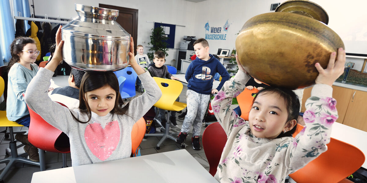 Zwei junge Mädchen sitzen an Tischen in einem Klassenzimmer und halten große Metalltöpfe auf ihren Köpfen, während andere Kinder im Hintergrund sitzen und zusehen.