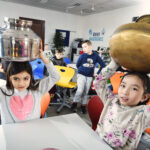 Zwei junge Mädchen sitzen an Tischen in einem Klassenzimmer und halten große Metalltöpfe auf ihren Köpfen, während andere Kinder im Hintergrund sitzen und zusehen.