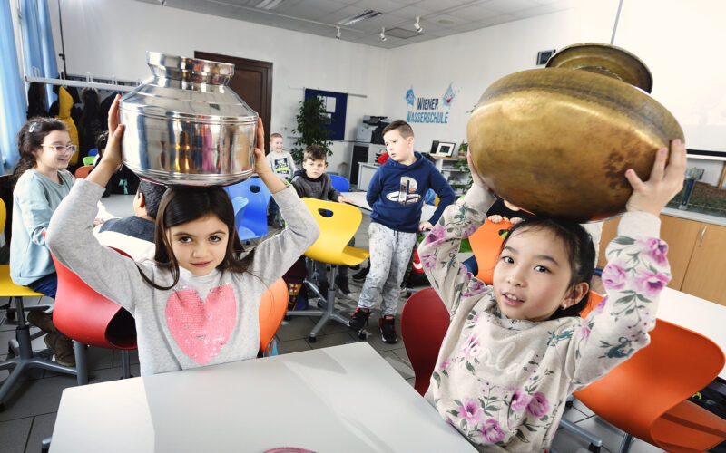 Zwei junge Mädchen sitzen an Tischen in einem Klassenzimmer und halten große Metalltöpfe auf ihren Köpfen, während andere Kinder im Hintergrund sitzen und zusehen.