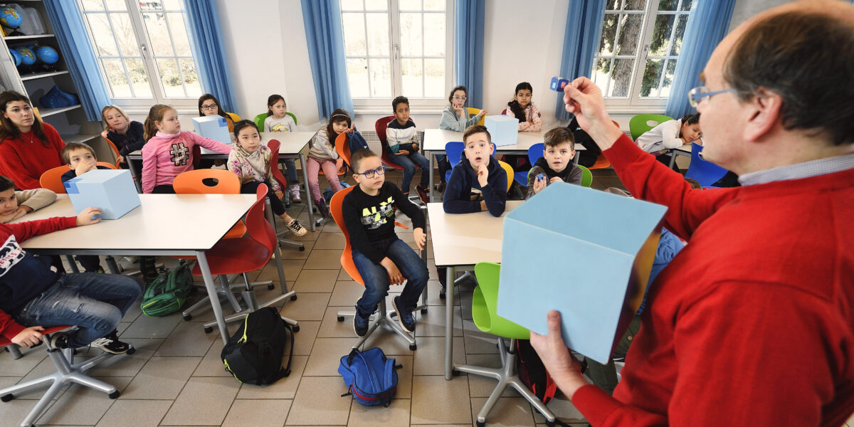 Eine Lehrerin in einem roten Pullover hält einen Gegenstand hoch, während eine Gruppe von Grundschülern in einem Klassenzimmer mit blauen Vorhängen und Tischen aufmerksam zusieht.