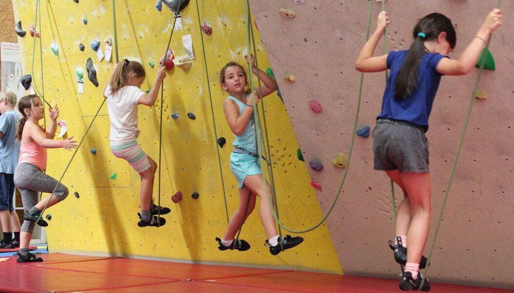 Vier Kinder in Klettergurten klettern mit Hilfe von Sicherungsseilen an einer Indoor-Felswand mit bunten Griffen. Sie tragen sportliche Kleidung und Kletterschuhe. Rote Sicherheitsmatten bedecken den Boden.