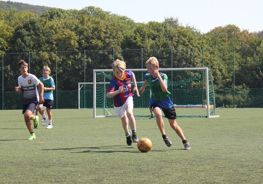 Zwei Jungen jagen einem Fußball auf einem Spielfeld hinterher, zwei weitere Jungen laufen hinter ihnen her und im Hintergrund sind Bäume zu sehen.