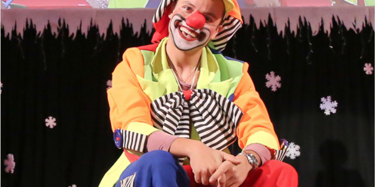 Eine als Clown verkleidete Person mit roter Nase, buntem Kostüm und Gesichtsbemalung sitzt auf der Bühne vor einem mit Schneeflocken dekorierten Hintergrund.
