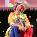 Eine als Clown verkleidete Person mit roter Nase, buntem Kostüm und Gesichtsbemalung sitzt auf der Bühne vor einem mit Schneeflocken dekorierten Hintergrund.
