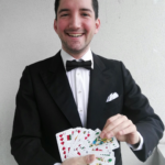 Ein Mann im Smoking und mit Fliege steht lächelnd vor einer schlichten Wand und hält ein aufgefächertes Kartenspiel in den Händen.