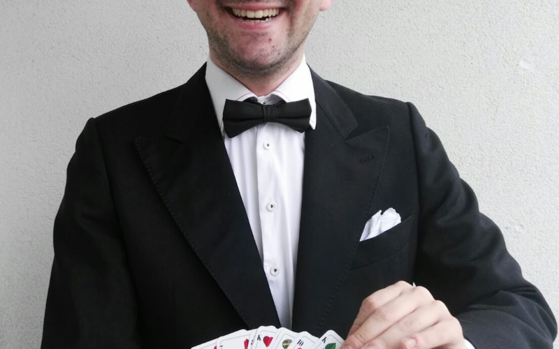 Ein Mann im Smoking und mit Fliege steht lächelnd vor einer schlichten Wand und hält ein aufgefächertes Kartenspiel in den Händen.