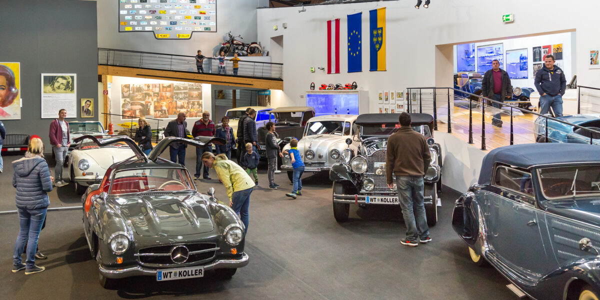 In einem gut beleuchteten Automobilmuseum werden Oldtimer ausgestellt, im Hintergrund sind mehrere Oldtimer, Fahnen und informative Exponate zu sehen.