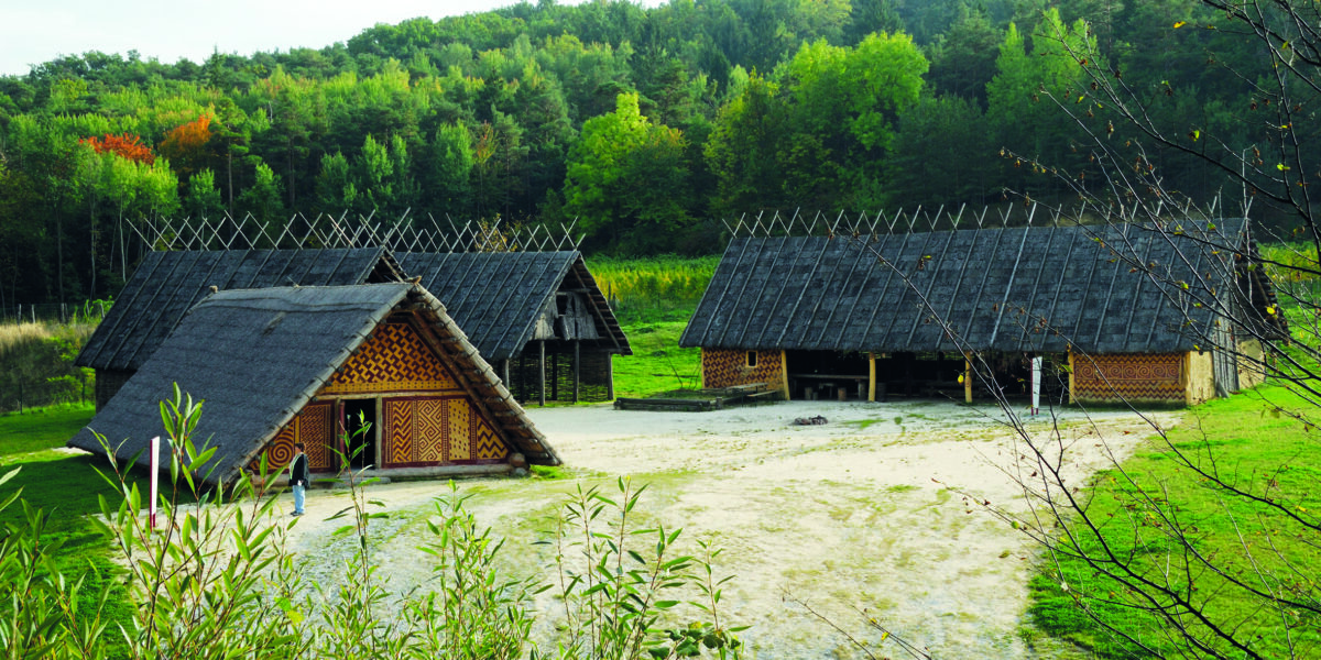 Traditionelle Holzbauten mit Strohdächern stehen auf einer gerodeten Fläche, umgeben von grünen Bäumen und grasbewachsenen Hügeln unter einem strahlenden Himmel.