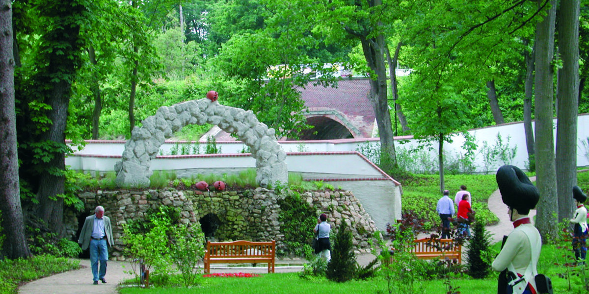 Die Besucher gehen durch einen Landschaftsgarten mit steinernen Torbögen, Bänken und gepflegten Blumenbeeten, die von hohen grünen Bäumen umgeben sind.
