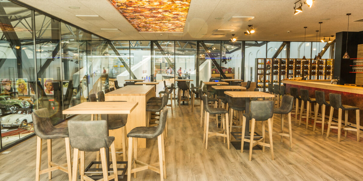 Modernes Café mit hohen Holztischen und -stühlen, großen Glasfenstern, Weinregalen und einer bunten Deckenverkleidung. Der Raum ist hell mit natürlichem Licht und zeitgenössischem Dekor.
