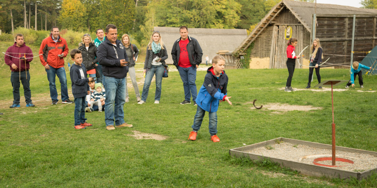 Eine Gruppe von Menschen sieht zu, wie ein Junge ein Hufeisen gegen einen Pfahl auf einer Wiese im Freien wirft, mit rustikalen Gebäuden im Hintergrund.