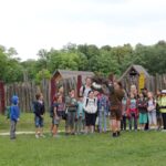 Eine Gruppe von Kindern und Erwachsenen steht draußen in der Nähe von Holzkonstruktionen und Bäumen und hört einer Person zu, die einen Greifvogel hält.