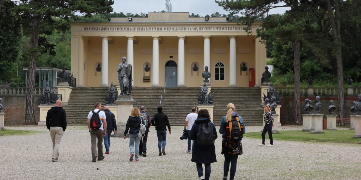 Eine Gruppe von Menschen geht auf ein gelbes neoklassizistisches Gebäude mit Säulen und Statuen davor zu, das von Bäumen umgeben ist.
