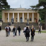 Eine Gruppe von Menschen geht auf ein gelbes neoklassizistisches Gebäude mit Säulen und Statuen davor zu, das von Bäumen umgeben ist.