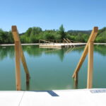 Ein Naturschwimmbad mit Holzgeländer und einer kleinen Holzplattform auf dem Wasser, umgeben von Grün unter einem klaren blauen Himmel.