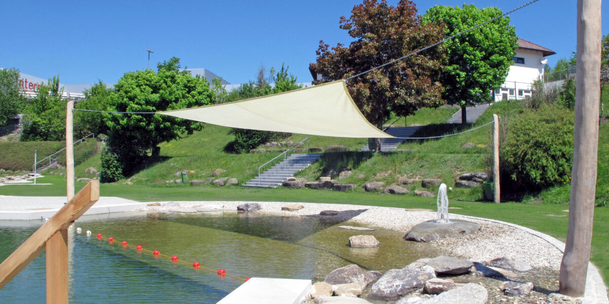 Ein natürlicher Schwimmteich im Freien mit einem Sonnensegel, Steinen, einem flachen Bereich zum Waten und orangefarbenen Bojen, umgeben von Gras und Bäumen unter einem klaren blauen Himmel.