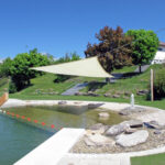 Ein natürlicher Schwimmteich im Freien mit einem Sonnensegel, Steinen, einem flachen Bereich zum Waten und orangefarbenen Bojen, umgeben von Gras und Bäumen unter einem klaren blauen Himmel.