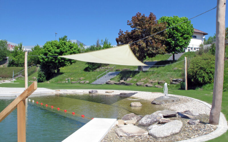Ein natürlicher Schwimmteich im Freien mit einem Sonnensegel, Steinen, einem flachen Bereich zum Waten und orangefarbenen Bojen, umgeben von Gras und Bäumen unter einem klaren blauen Himmel.