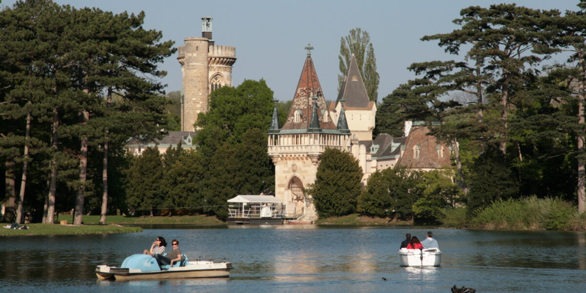 Tretbootfahrer auf einem ruhigen See mit einem historischen schlossähnlichen Gebäude und hohen Bäumen im Hintergrund unter einem klaren Himmel.