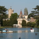 Tretbootfahrer auf einem ruhigen See mit einem historischen schlossähnlichen Gebäude und hohen Bäumen im Hintergrund unter einem klaren Himmel.