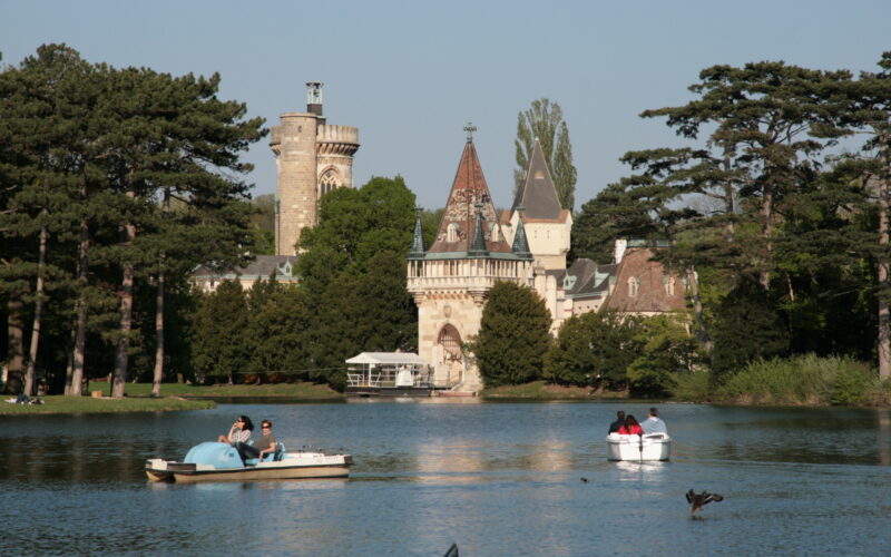 Tretbootfahrer auf einem ruhigen See mit einem historischen schlossähnlichen Gebäude und hohen Bäumen im Hintergrund unter einem klaren Himmel.
