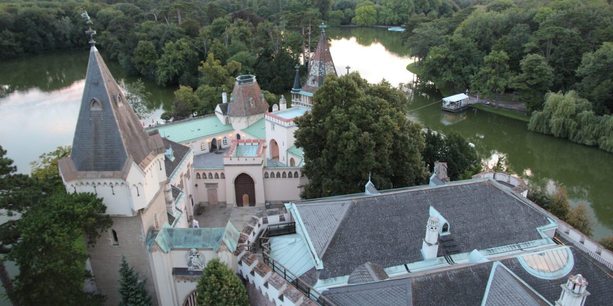 Luftaufnahme eines historischen Schlosses, umgeben von Bäumen und einem See, mit spitzen Türmen und Bergen im fernen Hintergrund.