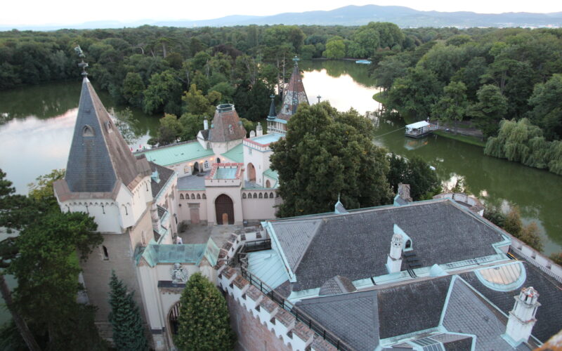 Luftaufnahme eines historischen Schlosses, umgeben von Bäumen und einem See, mit spitzen Türmen und Bergen im fernen Hintergrund.