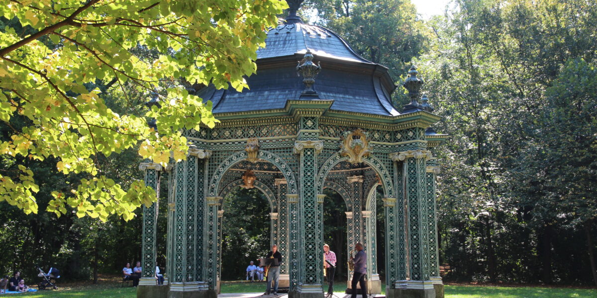 Ein verschnörkelter grüner Pavillon mit goldenen Akzenten steht in einem sonnenbeschienenen Park; mehrere Menschen sind darin und in der Nähe versammelt, umgeben von Bäumen und Gras.