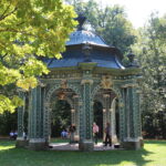 Ein verschnörkelter grüner Pavillon mit goldenen Akzenten steht in einem sonnenbeschienenen Park; mehrere Menschen sind darin und in der Nähe versammelt, umgeben von Bäumen und Gras.
