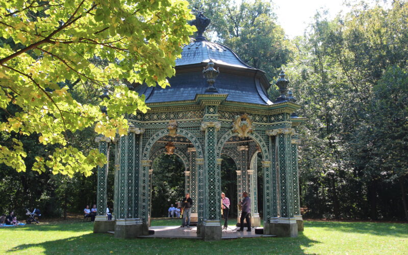 Ein verschnörkelter grüner Pavillon mit goldenen Akzenten steht in einem sonnenbeschienenen Park; mehrere Menschen sind darin und in der Nähe versammelt, umgeben von Bäumen und Gras.