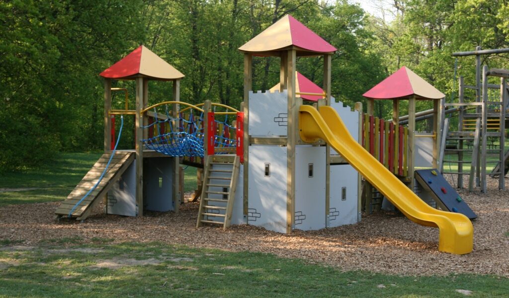 Spielplatz im Freien mit Rutschen, Kletterwänden und Brücken auf einer mit Mulch bedeckten Fläche, umgeben von Gras und Bäumen.