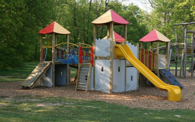Spielplatz im Freien mit Rutschen, Kletterwänden und Brücken auf einer mit Mulch bedeckten Fläche, umgeben von Gras und Bäumen.