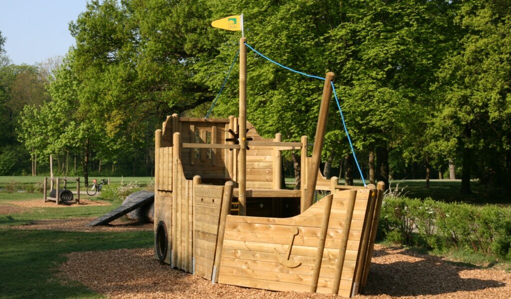 Ein hölzernes Spielgerät in Form eines Schiffes steht auf Mulch in einem Park, mit Bäumen und Gras im Hintergrund.