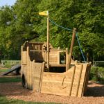 Ein hölzernes Spielgerät in Form eines Schiffes steht auf Mulch in einem Park, mit Bäumen und Gras im Hintergrund.