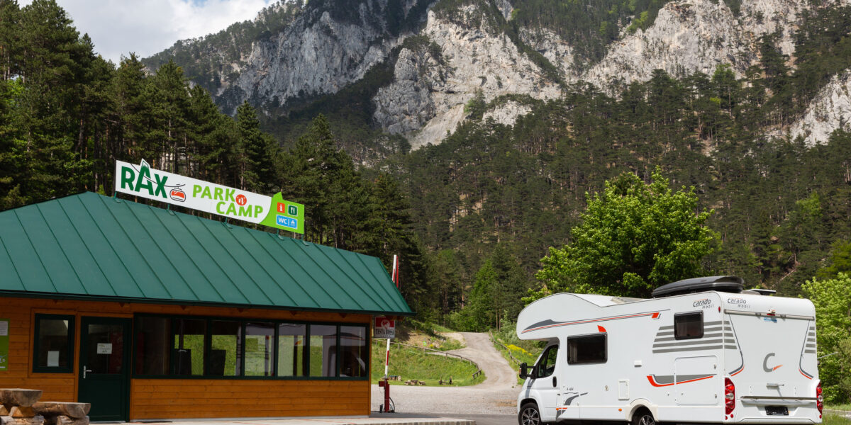 Ein weißes Wohnmobil steht in der Nähe des Eingangs zum Rax Park Camp, umgeben von Kiefern und Bergen unter einem teilweise bewölkten Himmel.