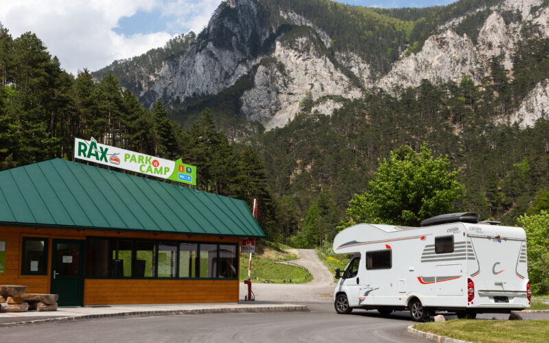 Ein weißes Wohnmobil steht in der Nähe des Eingangs zum Rax Park Camp, umgeben von Kiefern und Bergen unter einem teilweise bewölkten Himmel.