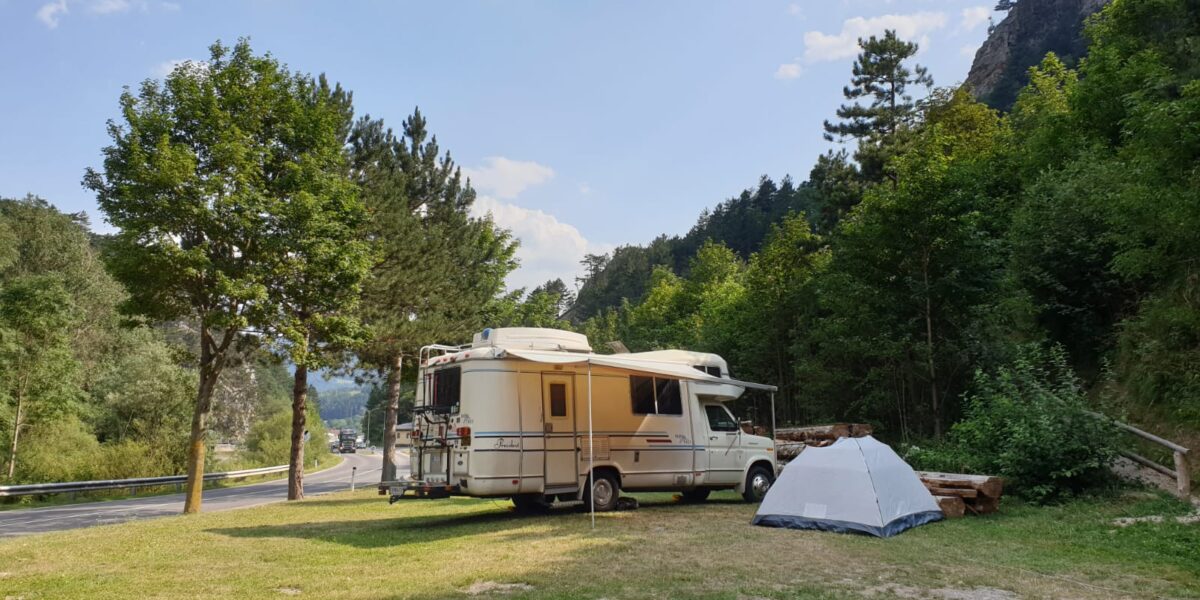 Ein weißes Wohnmobil und ein graues Zelt stehen auf einer Wiese in der Nähe einer Straße, umgeben von Bäumen und Hügeln unter einem teilweise bewölkten Himmel.