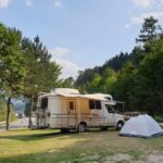Ein weißes Wohnmobil und ein graues Zelt stehen auf einer Wiese in der Nähe einer Straße, umgeben von Bäumen und Hügeln unter einem teilweise bewölkten Himmel.