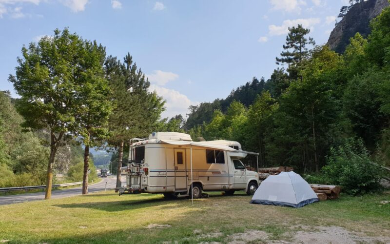 Ein weißes Wohnmobil und ein graues Zelt stehen auf einer Wiese in der Nähe einer Straße, umgeben von Bäumen und Hügeln unter einem teilweise bewölkten Himmel.