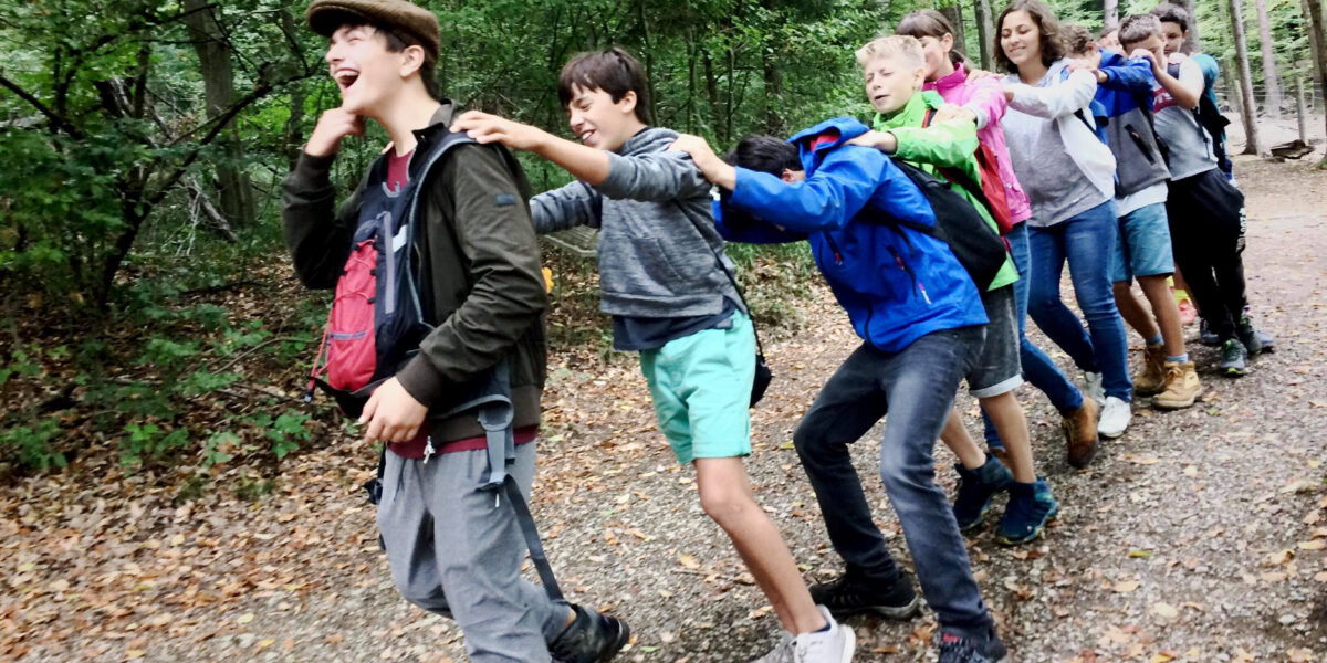 Eine Gruppe von Kindern läuft in einer Reihe durch ein Waldgebiet, wobei jedes Kind die Hände auf die Schultern des Vordermanns legt.