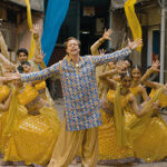 Eine Gruppe von Tänzern in gelber Tracht tritt hinter einer Person in blau-goldener Kleidung mit ausgestreckten Armen in einer mit blau-gelben Stoffen dekorierten Gasse auf.