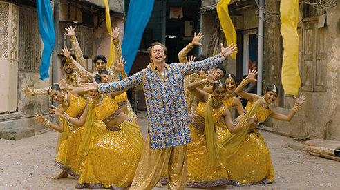 Eine Gruppe von Tänzern in gelber Tracht tritt hinter einer Person in blau-goldener Kleidung mit ausgestreckten Armen in einer mit blau-gelben Stoffen dekorierten Gasse auf.