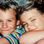 Zwei lächelnde, eng aneinander gelehnte Kinder schauen in die Kamera. Der Junge trägt ein gestreiftes Hemd und das Mädchen hat die Arme auf die Knie gestützt.