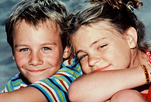Zwei lächelnde, eng aneinander gelehnte Kinder schauen in die Kamera. Der Junge trägt ein gestreiftes Hemd und das Mädchen hat die Arme auf die Knie gestützt.