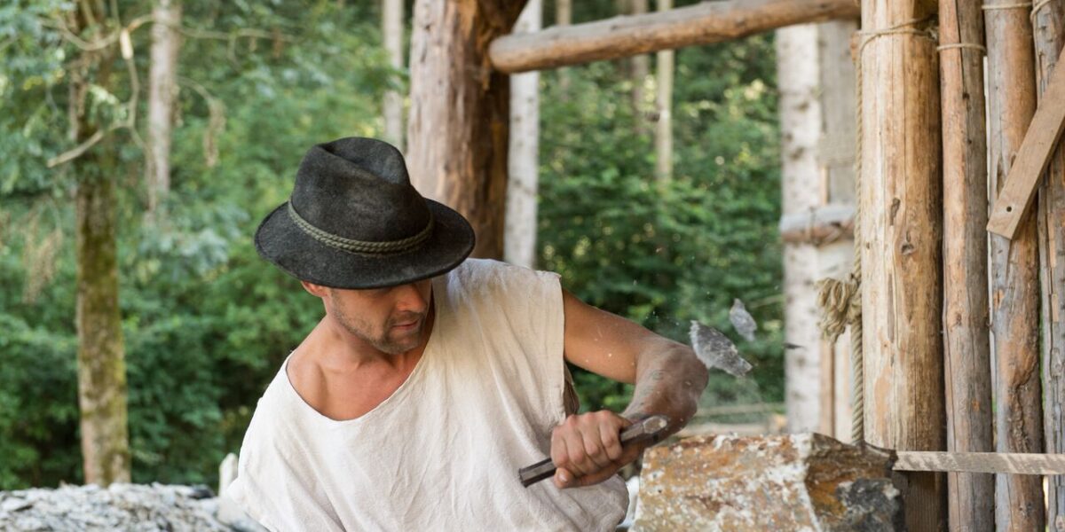 Ein Mann mit Hut und weißem Hemd bearbeitet einen Stein mit Hammer und Meißel in der Nähe einer Holzkonstruktion.